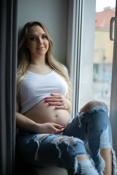 快乐的孕妇在白天用手放在窗台上摸着裸露的肚子 家休息 靠窗放松 年轻母亲的垂直家庭照片 怀孕和分娩 — 图库照片