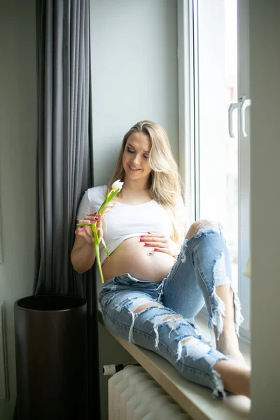快乐的孕妇在白天抱着新鲜的郁金香 在窗台上摸着裸露的肚子 家休息 靠窗放松 年轻母亲的垂直家庭照片 怀孕和分娩 — 图库照片