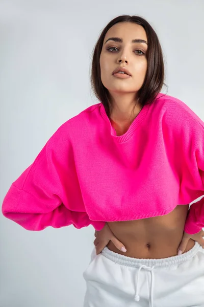 stock image Vertical dark haired confident, glorious woman with posh makeup in pink shirt with naked tummy, posing like fashion model with hands on hips in white studio. Sportswoman, healthy lifestyle, copy space