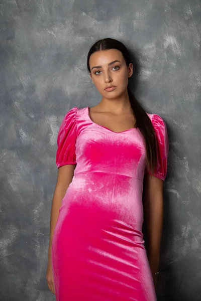Vertical calm, serious young woman with ponytail wear pink velvet dress, standing on gray studio background. Fashion industry, slim beauty body. Presenting modern outfit and textile. Copy space