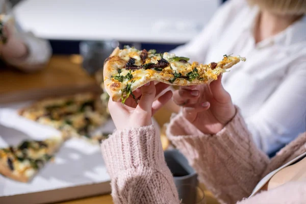 Cropped photo of woman sitting at table among friends relatives , holding piece of vegetable pizza, enjoying food, relaxing at home. Celebration, birthday, fast food, party, togetherness. Soft focus.