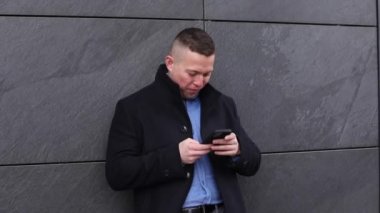 İyi kesilmiş mavi kazaklı, siyah ceketli yakışıklı iş adamının videosu. Gri kiremit duvara karşı ofis merkezinin dışında gülümseyen yeni telefon daktilosuna bakıyor. Yaşam biçimi