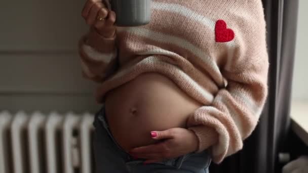 認識できない若い妊娠中の女性は 自宅に立って ジーンズのアプリケーションでベージュのセーターを着て コーヒー茶のカップを保持し 飲んで 裸の腹を撫でています — ストック動画