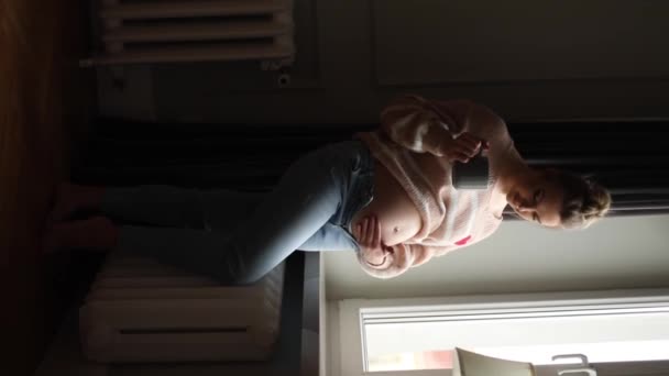 漂亮的年轻快乐的孕妇穿着米黄色毛衣 牛仔裤 站在窗前 爱抚赤身裸体 享受怀孕的快乐 — 图库视频影像