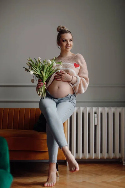 穿着粉色针织套头衫和牛仔裤 赤脚坐在沙发上 束着郁金香的年轻孕妇的画像 她赤脚着地 梦幻般地笑着 搂着她的胃 — 图库照片