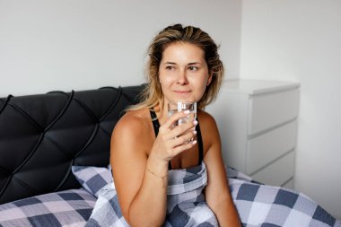 Mutlu kadın sabah uyanır ve yatakta oturup su içer. Düzgün beslenme. Sağlıklı bir yaşam tarzı. Olumlu duygular. Su dengesi. Güne iyi bir başlangıç. Diyet ve kilo kaybı. Güzellik ve sağlık. 