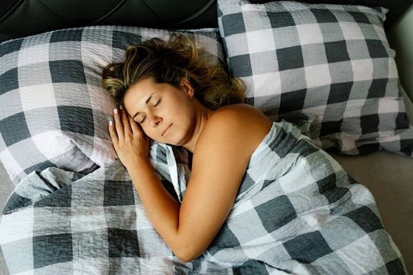 Charming tanned woman sleeps in bed, turning on her side and putting her hand under her cheek. View from above. Rest and healthy sleep. Calm and serenity. Healthy lifestyle. Natural materials.