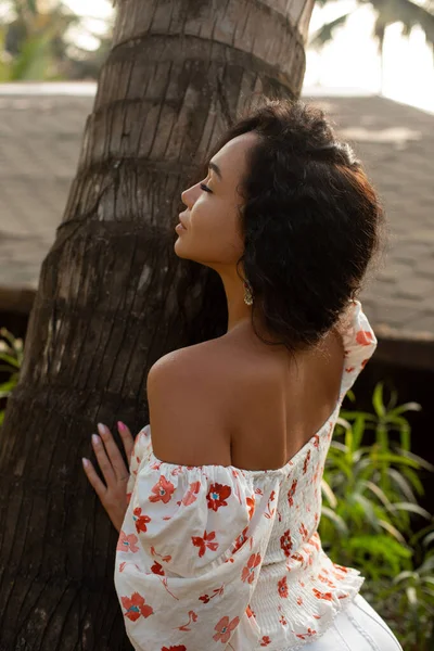 Rear view. Beautiful line of female shoulders. Young tanned woman in open-shouldered blouse with her eyes closed catches rays of sun leaning against tree. Tourism and travel. Skin care. Enjoying.