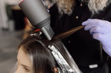 Güzellik salonunda oturan genç bir kadının yan görüntüsü. Tanımlanamayan kadın kuaför boyacısı. Uzun saç telini kurutmak için mavi lastik eldivenli kurutucu kullanıyor. Saç boyama, renklendirme, dengeleme.