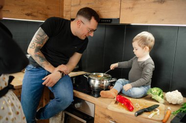 Küçük bir çocuğun aile portresi, mutfakta yemek yapan genç bir adam. Bebek bebek babasıyla birlikte tencerenin yanındaki masada oturuyor, sebze çorbasını çorba kaşığıyla karıştırıyor, annesi izliyor, oğlunu kontrol ediyor..