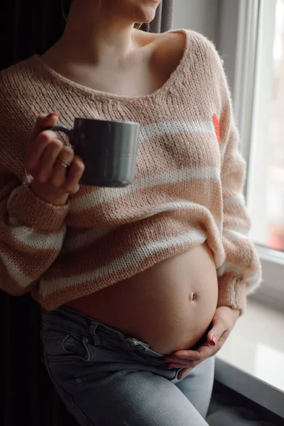 Cropped photo of young pregnant woman wearing beige sweater, jeans, standing at window, holding cup, caressing touching naked belly. Motherhood, pregnancy, maternity, expectation, health. Vertical.