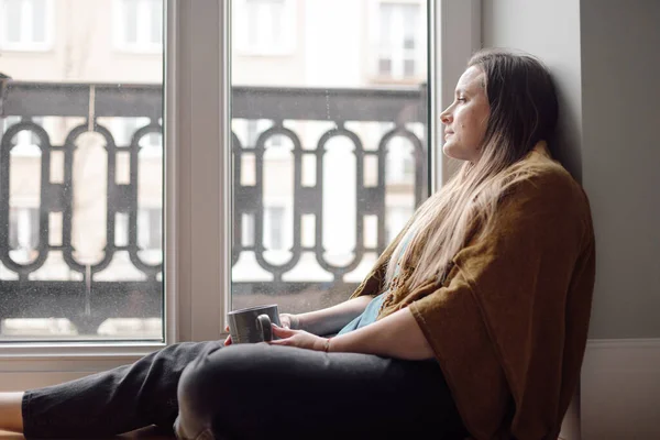 Portrait of middle-aged depressed woman with long hair sit on floor, looking through window, holding grey cup, suffering from psychological problems, thinking. Stress, depression, negative emotions.