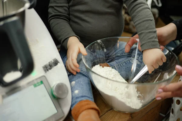Cropped photo of family of little boy and young woman cooking baking in kitchen at home. Baby toddler sitting on table near kneader cooking machine, filling spoon with flour, mother holding bowl.