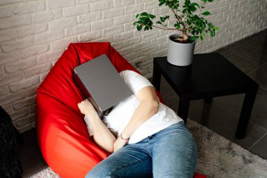 Yukarıdan bak. Beyaz tişörtlü ve kot pantolonlu tanınmayan bir kadın evinde turuncu koltukta otururken dizüstü bilgisayarla uyuyakaldı. İnternette evde çalışıyorum. Yorgunluk ve tükenmişlik. Bağımsız.