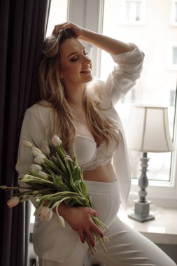 Kapalı gözlü, beyaz takım elbiseli, dantelli sutyen giyen, pencerede duran, elinde buket dolusu beyaz lale tutan, uzun sarı saçlı hamile bir kadının yan görüntüsü. Annelik, beklenti. 