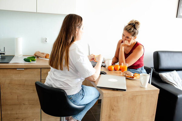 Communication and mutual understanding. Two cute girlfriends are sitting facing each other in kitchen talking, having lunch with fruit, laptop is lying next to them. Comfort and work. Home office,.