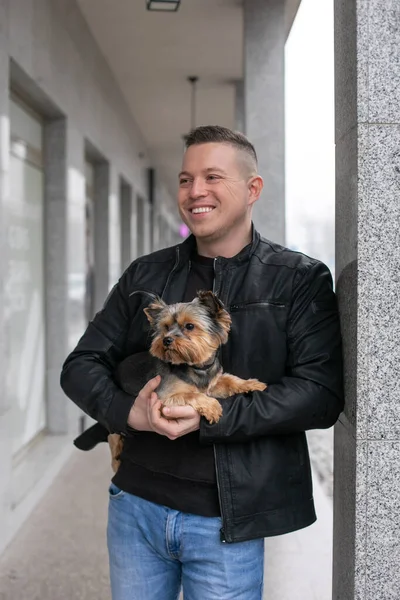 Portrait of clever young well-cut businessman in jeans and black sneakers black sweater black jacket smiling laughing at and staying with dogs on leash outside the office in urban city