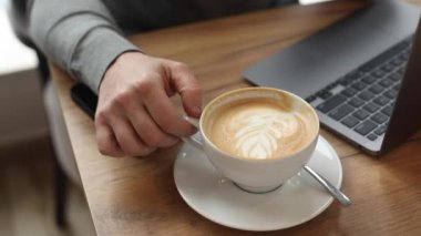 Gri kazak giyen, kremalı kahve içen, çay kaşığı beyaz tabakta, ahşap masadaki pahalı gri Macbook 'un yanında iş arkadaşlarını bekleyen sportif iş adamının videosu.