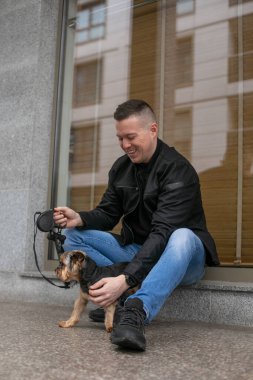 Kot pantolon ve siyah spor ayakkabılı genç iş adamının portresi siyah kazak siyah ceket gülüyor ve şehir merkezindeki ofisin dışında tasmalı köpeklerle oturuyor.