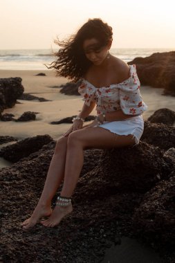 Dikey nazik, yalın ayaklı, uzun saçlı, esinti saçı olan, açık yaz elbiseli, deniz kıyısındaki kaya taşlarının üzerinde oturan, yaya olarak aşağıya bakan kadın. Seyahat, yaz tatili macerası