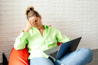 Baş ağrısı ve yorgunluk. Açık yeşil tişörtlü ve kot pantolonlu genç bir kadın evinde dizüstü bilgisayarla oturmaktan rahatsız oluyor. Yorgunluk ve tükenmişlik. Serbest çalışıyorum. Modern teknolojiler. 