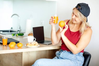 Beyzbol şapkalı ve tişörtlü alımlı sarışın mutfağında kahvaltı yapıyor taze sıkılmış portakal suyu ve portakal suyunun rengini karşılaştırıyor. Evde online çalışıyor. Sağlıklı beslenme. Mizah. 