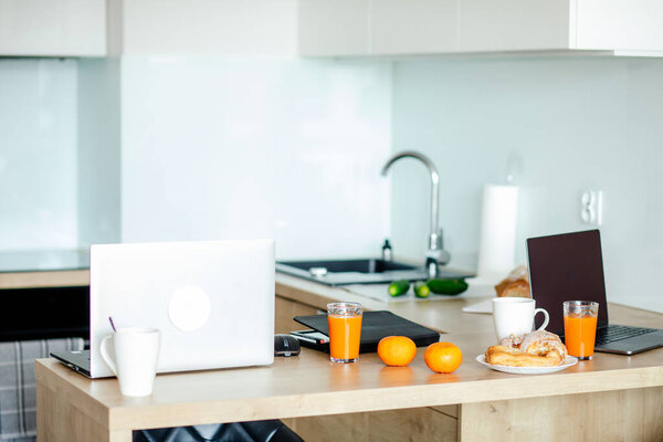 Modern kitchen without people with wooden work surface, which is used as home office, there are two laptops on it, there are oranges, there are coffee cups and glasses with juice. Home office. 