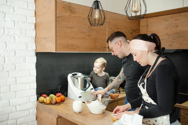 Portrait of family of young woman, little boy and man baking in kitchen at home. Son sitting on table near kneader cooking machine, father turning on device, tapping on display. Relationship, pastry.