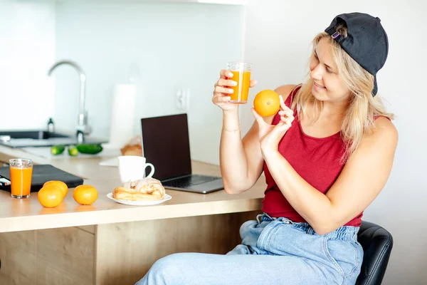 Charming blonde in baseball cap and T-shirt is having breakfast in her kitchen, comparing color of freshly squeezed orange juice and orange itself. Works online at home. Healthy eating. Humor.