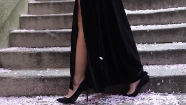 外の風の強い天候で階段の近くを歩く女性のハイヒールビジネスシューズ黒のドレスのビデオ 顔は見えない 美脚だ 階段の上にスタイリッシュなアパレルを持つ豪華な女性 — ストック動画