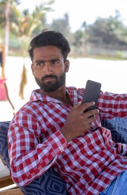 Yatılı adamın portresi. Gömlekli, kot pantolonlu, kalın saçlı, yan yana oturan ve telefon kullanarak kumsalda, gri ağaçlı bir plajda yapılan ayinler sırasında arama ve arama yapan. 