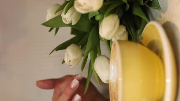詳細を動画で確認できませんが 女性は美しい花束に触れていますヌードマニキュア黄色のカップの近くにクリームコーヒーミルクと白いテーブルの上にカフェ内 — ストック動画