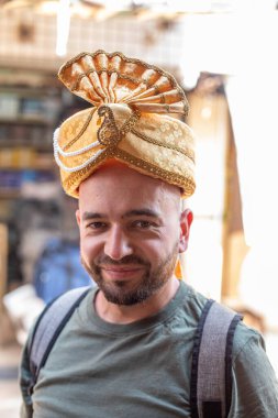 Haki tişört giyen gülümseyen sakallı genç bir adamın portresi, damat için sarı sarı Hint şapkası türbanı, sırt çantası, dışarıda dikilen. Seyahat, turizm, tatil, tatil, yaz.