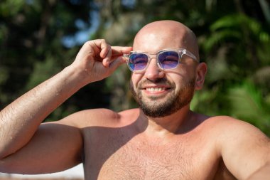 Güneşli bir günde palmiye ağaçlarının yanında oturan, rahatlatıcı poz veren, mavi güneş gözlüklerine dokunan mutlu kel sakallı adamın portresi. Seyahat, turizm, tatil, yaz, tatil, tatil.