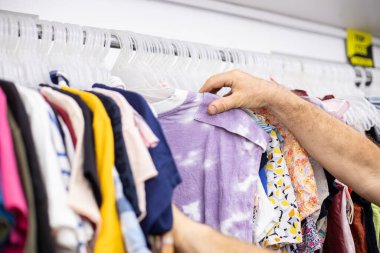 Tanınmayan bir adam elbise mağazasında asılı duran yazlık pamuk tişörtü seçiyor. Geniş bir kıyafet seçimi. Küçük bir iş. Yetişkin kıyafetleri. Parlak kıyafetler. Çıkış noktası. 