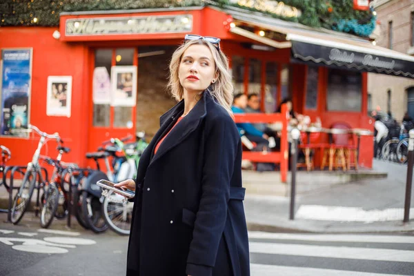 stock image Adorable young girl with white, wavy, flowing hair, in black coat look back while walking on streets of Paris. Confident lady with red lipstick traveling in sunny France near cafe.