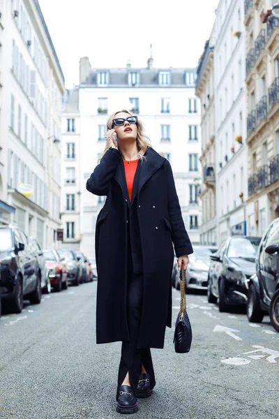 Charming lady with blond hair wearing sunglasses and black coat talking on phone while looking up at sky outdoor. Adult woman walking in Paris near huge buildings.