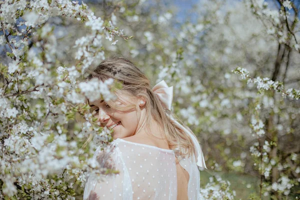 身着婚纱的优雅女士站在樱花环抱下 带着柔和的微笑回头看 年轻的新娘 头戴大蝴蝶结 在樱花树旁散步 享受着阳光灿烂的日子 — 图库照片