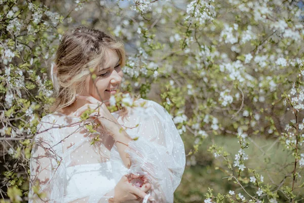 年轻的新娘笑着欣赏大自然 同时站在樱花枝旁 漂亮的女士穿着婚纱 身边环绕着白樱花 在阳光灿烂的夏日呆在户外 — 图库照片