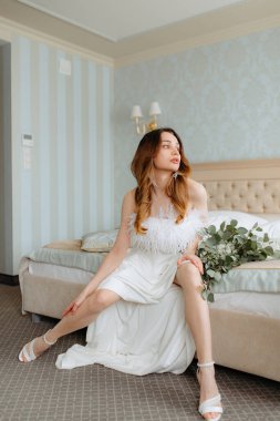 Yatağın kenarına tüylerle süslenmiş, öne eğilip yana bakan muhteşem gelinlikli kadın. Çiçek buketi kızıl kahverengi saçlı çekici bayanın yanında yatıyor..