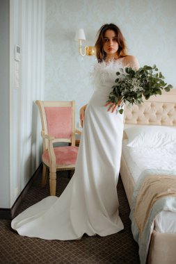 Uzun omuzsuz bir düğün elbisesi giymiş, yatak odasında yatağın yanında duran tüylerle süslenmiş hoş bir genç kadın. Koyu saçlı kadın elinde bir buket düğün çiçeği tutuyor..