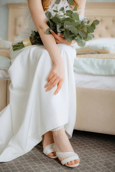 Unrecognizable young woman sitting on bed in luxurious long wedding gown with feathers and white high heels shoes decorated with pearls. Bride with red and brown hair hold bouquet of blossoms on lap.