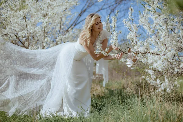 穿着婚纱 可敬的年轻女子站在樱花旁 倚在树枝上笑着 女士的高跟鞋挂在草裙鞋上 新娘要穿上鞋子 — 图库照片