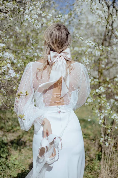 年轻的女人穿着婚纱 背带着绳子 站在樱桃树中间 手里拿着一双白高跟鞋 金发碧眼的新娘的后视镜 垂直方向 — 图库照片