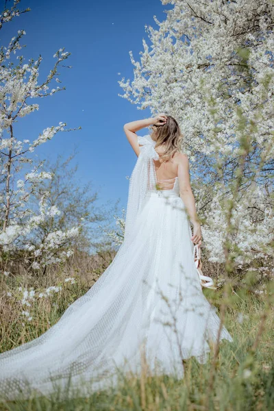 美丽的金发碧眼的新娘穿着长长的婚纱 背带着丝带 站在白樱花旁 手里拿着一双新娘高跟鞋 垂直方向 — 图库照片
