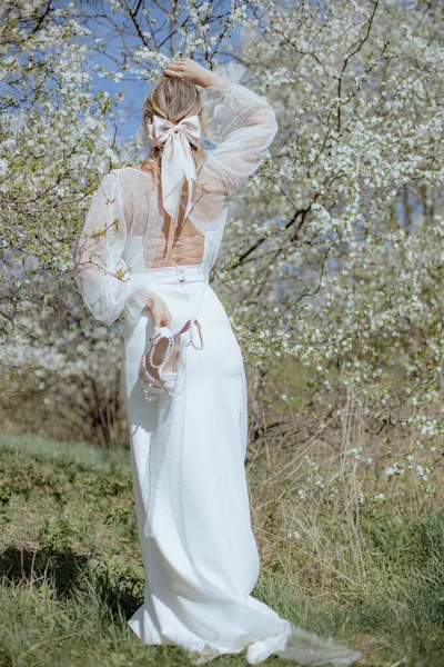 桜の花の間に立って 真珠で飾られた白いハイヒールの靴のペアを保持し 背面に文字列を持つ長い結婚式のガウンを着て髪に弓を持つ若い女性のリアビュー — ストック写真