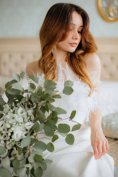 ふわふわの羽がベッドの上に座り 白い花の花束を手にして飾られた肩のない結婚式の衣装を身に着けている豪華な女性 若いですブルネット持ち上げる生地のドレスと下を見る — ストック写真