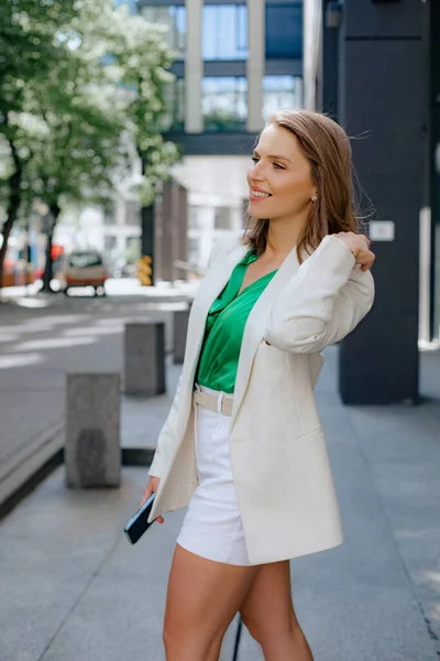 Tanned, confident business lady in elegant jacket walks down street of city to business meeting with phone in her hand. Success and career. Own business and entrepreneurship. Modern business women.