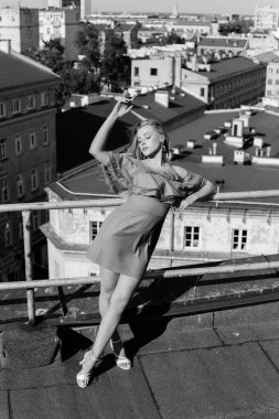 Kabarık elbiseli ve ince topuklu ayakkabılı genç bayan yüksek katlı bir binanın çatısında dikiliyor ve kapalı gözlerle çitlere yaslanıyor. Kadın kolunu kaldırıyor ve temiz havanın tadını çıkarıyor. Dikey. Siyah ve beyaz.