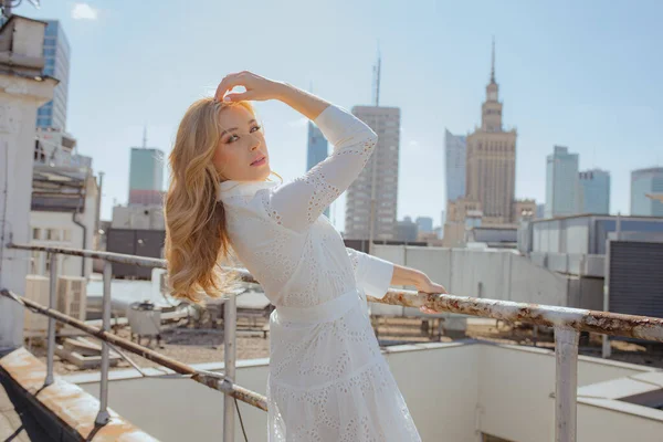 背中を傾け ブロンドの髪に触れながら手でフェンスに保持刺繍と白いドレスの楽しい女性 都会を背景に高層ビルの屋上に立つ上品な女性 — ストック写真